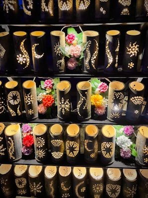 A display of bamboo lanterns with intricate patterns carved into their surfaces, illuminated from within. The lanterns are arranged in rows and accompanied by colorful artificial flowers interspersed among them.