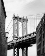 A black and white photograph of a large suspension bridge seen from between two tall buildings. The bridge features intricate steel beams and cables, with an imposing structure that highlights its engineering marvel. The sky is overcast, adding depth to the monochromatic scene.