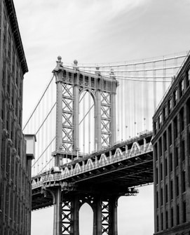 A black and white photograph of a large suspension bridge seen from between two tall buildings. The bridge features intricate steel beams and cables, with an imposing structure that highlights its engineering marvel. The sky is overcast, adding depth to the monochromatic scene.