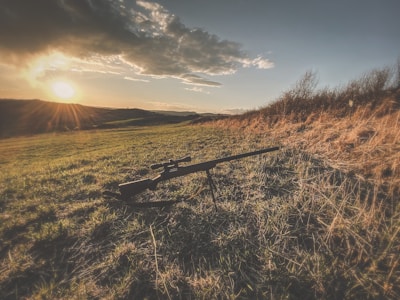 A long rifle with a scope is lying on the ground of a grassy field under a setting sun. The sky is partly cloudy, and the landscape features hills and dry grass.