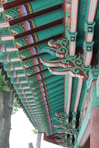 Detailed architectural elements of a traditional wooden structure, featuring ornate and colorful patterns. The beams are painted with intricate designs in shades of green, red, and blue, highlighting the craftsmanship involved. Natural wood grain is visible beneath the paint, adding texture to the design. The setting includes trees in the background, contributing to an outdoor environment.