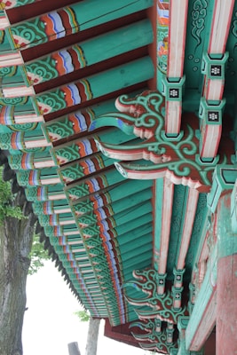 Detailed architectural elements of a traditional wooden structure, featuring ornate and colorful patterns. The beams are painted with intricate designs in shades of green, red, and blue, highlighting the craftsmanship involved. Natural wood grain is visible beneath the paint, adding texture to the design. The setting includes trees in the background, contributing to an outdoor environment.