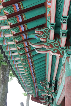 Detailed architectural elements of a traditional wooden structure, featuring ornate and colorful patterns. The beams are painted with intricate designs in shades of green, red, and blue, highlighting the craftsmanship involved. Natural wood grain is visible beneath the paint, adding texture to the design. The setting includes trees in the background, contributing to an outdoor environment.