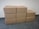 Eight cardboard boxes arranged in a staggered manner on a tiled floor against a textured white wall. Four boxes form the bottom row, three boxes form the middle row, and one box sits at the top of the arrangement.