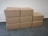 Eight cardboard boxes arranged in a staggered manner on a tiled floor against a textured white wall. Four boxes form the bottom row, three boxes form the middle row, and one box sits at the top of the arrangement.