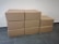 Eight cardboard boxes arranged in a staggered manner on a tiled floor against a textured white wall. Four boxes form the bottom row, three boxes form the middle row, and one box sits at the top of the arrangement.