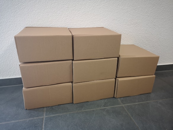 Eight cardboard boxes arranged in a staggered manner on a tiled floor against a textured white wall. Four boxes form the bottom row, three boxes form the middle row, and one box sits at the top of the arrangement.