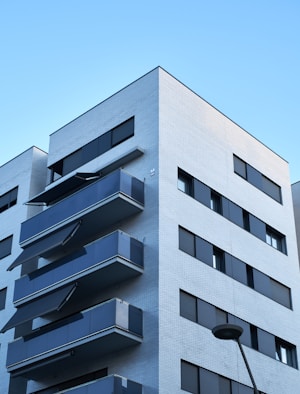 Modern apartment building with multiple balconies on each floor. The structure features light-colored brickwork and large, dark glass windows. A streetlight is visible in the foreground, with a clear blue sky in the background.