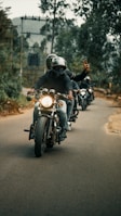 A group of motorcyclists riding down a tree-lined road, with the lead rider showing a peace sign. The backdrop includes blurred trees and overcast skies, creating a sense of movement and camaraderie.