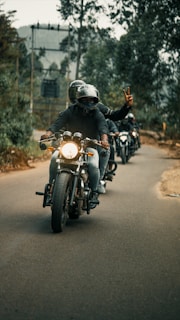 A group of motorcyclists riding down a tree-lined road, with the lead rider showing a peace sign. The backdrop includes blurred trees and overcast skies, creating a sense of movement and camaraderie.