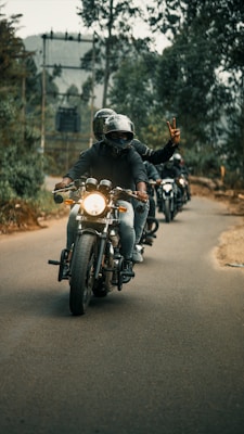 A group of motorcyclists riding down a tree-lined road, with the lead rider showing a peace sign. The backdrop includes blurred trees and overcast skies, creating a sense of movement and camaraderie.