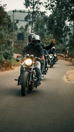 A group of motorcyclists riding down a tree-lined road, with the lead rider showing a peace sign. The backdrop includes blurred trees and overcast skies, creating a sense of movement and camaraderie.