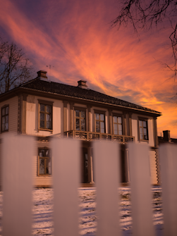 A two-story wooden house with a warm, inviting exterior sits behind a white picket fence. The sunset sky above is painted with vibrant pink and orange hues, casting a serene glow over the snowy ground. Bare tree branches frame the scene, adding a touch of winter charm.