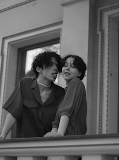 Two people are standing on a balcony. One person is playfully leaning towards the other, creating a sense of intimacy and affection. They both appear relaxed and are wearing casual clothing. The setting is sophisticated, with ornate architectural details visible in the background. The image is in black and white, adding a classic and timeless feel.