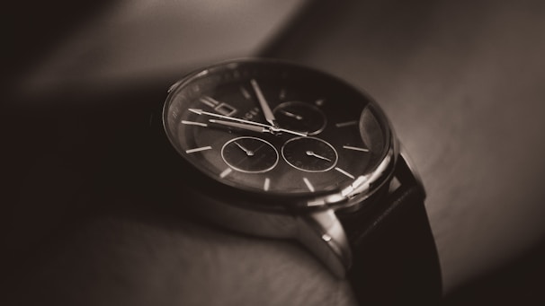 A close-up of a stylish wristwatch with a black leather band and a dark, sleek watch face featuring multiple dials and slim metal hands.