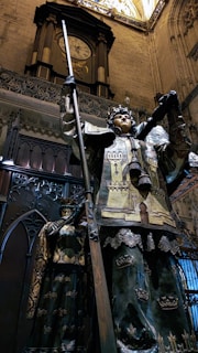 A grand and intricate statue of a warrior figure clad in ornate armor, holding a staff or spear, set against the backdrop of an elaborate historical interior with a large clock above. The figure features detailed designs, including crowns and patterns, with a dark patina suggesting age.