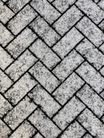 A close-up view of a textured surface made up of interlocking rectangular concrete pavers arranged in a herringbone pattern. The rough, gritty texture of the pavers is visible, with small stones embedded in the surface.