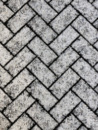 A close-up view of a textured surface made up of interlocking rectangular concrete pavers arranged in a herringbone pattern. The rough, gritty texture of the pavers is visible, with small stones embedded in the surface.