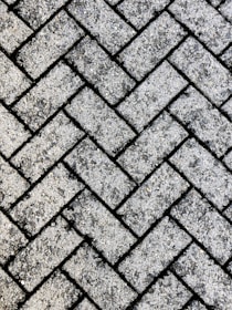 A close-up view of a textured surface made up of interlocking rectangular concrete pavers arranged in a herringbone pattern. The rough, gritty texture of the pavers is visible, with small stones embedded in the surface.