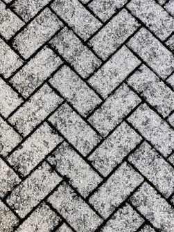 A close-up view of a textured surface made up of interlocking rectangular concrete pavers arranged in a herringbone pattern. The rough, gritty texture of the pavers is visible, with small stones embedded in the surface.