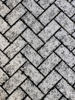 A close-up view of a textured surface made up of interlocking rectangular concrete pavers arranged in a herringbone pattern. The rough, gritty texture of the pavers is visible, with small stones embedded in the surface.