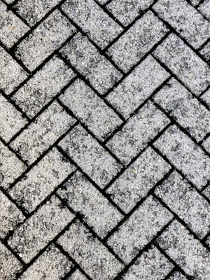 A close-up view of a textured surface made up of interlocking rectangular concrete pavers arranged in a herringbone pattern. The rough, gritty texture of the pavers is visible, with small stones embedded in the surface.