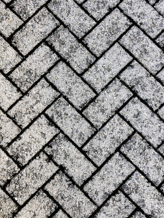 A close-up view of a textured surface made up of interlocking rectangular concrete pavers arranged in a herringbone pattern. The rough, gritty texture of the pavers is visible, with small stones embedded in the surface.