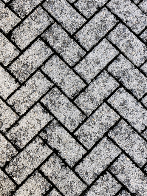 A close-up view of a textured surface made up of interlocking rectangular concrete pavers arranged in a herringbone pattern. The rough, gritty texture of the pavers is visible, with small stones embedded in the surface.