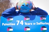 A person wearing a globe mask with a sad expression. The individual is holding a sign that reads 'Palestine 74 Years of Suffering' with Palestinian flags displayed repetitively. The person is dressed in blue.