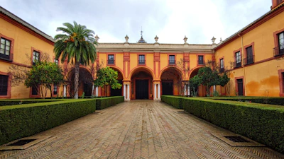 A symmetrical courtyard features a central pathway lined with manicured hedges leading to an archway. The surrounding buildings are adorned with a warm, earthy hue and classic architectural elements. Leafy trees and a prominent palm tree enhance the area's serene atmosphere.