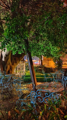 An outdoor setting with ornate blue metal chairs and tables arranged on a wooden deck. A large tree with lush green foliage and a trunk is dominant in the scene. Decorative lights hang from the tree, creating a warm ambiance. The background shows blurred lights from a building or structure, contributing to a cozy atmosphere.