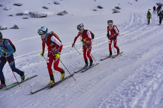 A group of cross-country skiers in red and blue outfits are skiing uphill on a snowy landscape. They are equipped with helmets, goggles, and backpacks, and holding ski poles. The snow-covered ground is surrounded by scattered rocks.