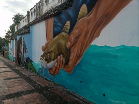 A large mural painted on a street wall displays a pair of hands holding a fish against a backdrop of water. The mural features vibrant colors and displays a realistic style, capturing the drape and texture of both the hands and the fish. The wall continues with a faded, historic building in the background, overgrown with some foliage.