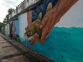 A large mural painted on a street wall displays a pair of hands holding a fish against a backdrop of water. The mural features vibrant colors and displays a realistic style, capturing the drape and texture of both the hands and the fish. The wall continues with a faded, historic building in the background, overgrown with some foliage.