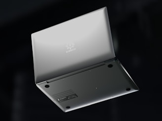 A silver laptop is shown upside down with its lid partially open, revealing the bottom side. The brand name 'Slimbook' is visible on the exterior, with rubber feet and ventilation details on the base. The background is dark, contrasting with the laptop's sleek metallic appearance.