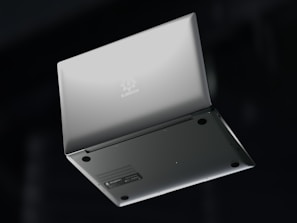A silver laptop is shown upside down with its lid partially open, revealing the bottom side. The brand name 'Slimbook' is visible on the exterior, with rubber feet and ventilation details on the base. The background is dark, contrasting with the laptop's sleek metallic appearance.