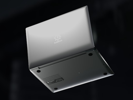 A silver laptop is shown upside down with its lid partially open, revealing the bottom side. The brand name 'Slimbook' is visible on the exterior, with rubber feet and ventilation details on the base. The background is dark, contrasting with the laptop's sleek metallic appearance.