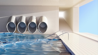 A clean, tiled indoor pool area with four large, cylindrical pipes on the wall. The pool water reflects light, creating dynamic patterns. A ramp with handrails leads down into the water, and there's a bright, rectangular passageway leading outside.