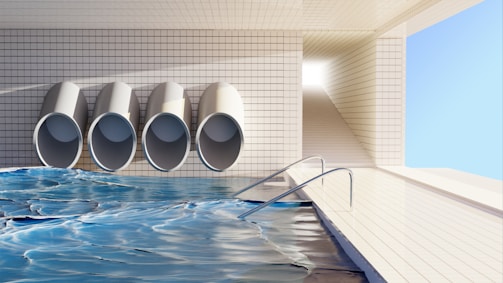 A clean, tiled indoor pool area with four large, cylindrical pipes on the wall. The pool water reflects light, creating dynamic patterns. A ramp with handrails leads down into the water, and there's a bright, rectangular passageway leading outside.