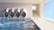 A clean, tiled indoor pool area with four large, cylindrical pipes on the wall. The pool water reflects light, creating dynamic patterns. A ramp with handrails leads down into the water, and there's a bright, rectangular passageway leading outside.