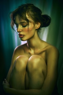 A serene woman with closed eyes is seated in a contemplative pose, hugging her knees. The soft lighting creates a gentle play of shadows and highlights on her skin. Her hair is styled in a loose bun, and her makeup is subtle yet elegant, with a focus on her eyeshadow. The background is a gradient of deep greens and blues, adding to the image's calm and introspective mood.