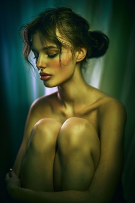 A serene woman with closed eyes is seated in a contemplative pose, hugging her knees. The soft lighting creates a gentle play of shadows and highlights on her skin. Her hair is styled in a loose bun, and her makeup is subtle yet elegant, with a focus on her eyeshadow. The background is a gradient of deep greens and blues, adding to the image's calm and introspective mood.