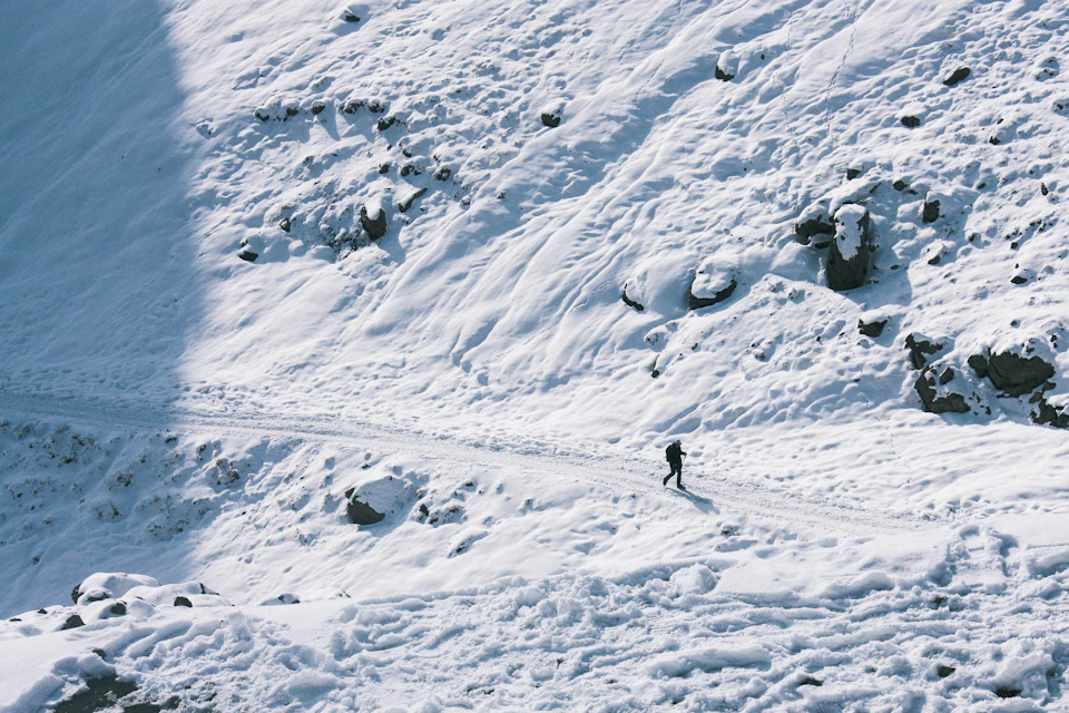 The image depicts a lone figure skiing down a snowy mountain slope, surrounded by large rocks and boulders on both sides of the trail. The skier is positioned centrally in the frame, moving towards the right side of the image. The scene conveys a sense of solitude and adventure, with the skier carving their path through the untouched snow.

The sky above is clear blue, providing a beautiful backdrop to the winter landscape below.