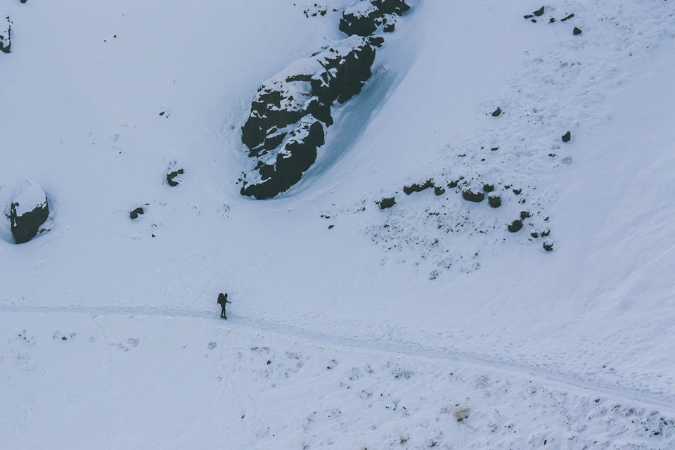 The image depicts a lone hiker traversing a snowy mountain trail, surrounded by large rocks and boulders. The hiker is wearing a backpack and appears to be moving away from the camera towards the right side of the frame. The trail winds through the snow-covered landscape, with patches of grass visible in the distance.