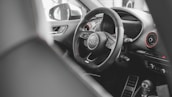 A modern car interior featuring a detailed view of a steering wheel with the Audi logo prominently displayed. The dashboard includes visible air vents with red accents and various controls, while the gear shift lever is partly visible in the foreground.