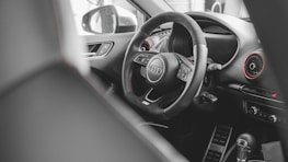A modern car interior featuring a detailed view of a steering wheel with the Audi logo prominently displayed. The dashboard includes visible air vents with red accents and various controls, while the gear shift lever is partly visible in the foreground.
