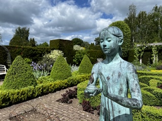 A serene garden scene with meticulously shaped shrubs, vibrant flower beds, and a large bronze statue of a young person holding a small object, possibly a flame or flower. The garden is framed by lush greenery and a cloudy blue sky, creating a peaceful and natural setting.