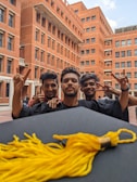 Three young men in graduation gowns are celebrating in a courtyard with brick buildings in the background. They are smiling and holding up peace signs. A graduation cap with a gold tassel is in the foreground.