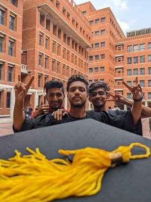 Three young men in graduation gowns are celebrating in a courtyard with brick buildings in the background. They are smiling and holding up peace signs. A graduation cap with a gold tassel is in the foreground.