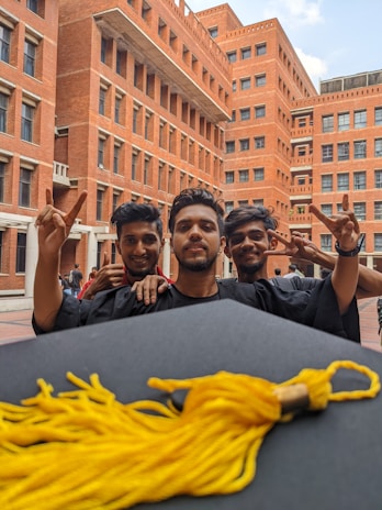 Three young men in graduation gowns are celebrating in a courtyard with brick buildings in the background. They are smiling and holding up peace signs. A graduation cap with a gold tassel is in the foreground.